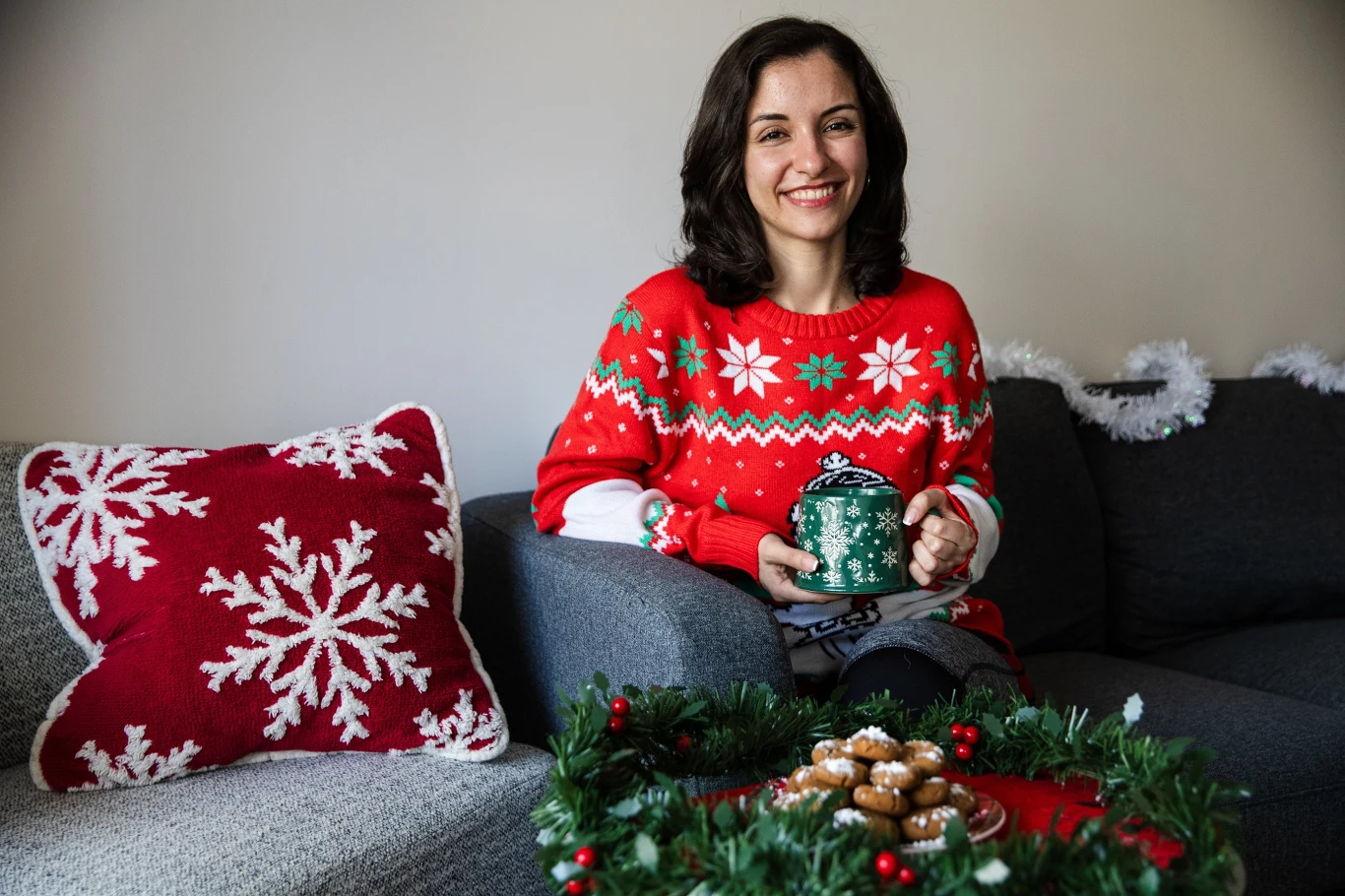 Świąteczny dress code Uśmiechnięta kobieta w czerwonym swetrze świątecznym siedzi na szarej sofie, trzyma kubek w dłoniach, obok leży poduszka z motywem śnieżynek oraz stroik z ciasteczkami.