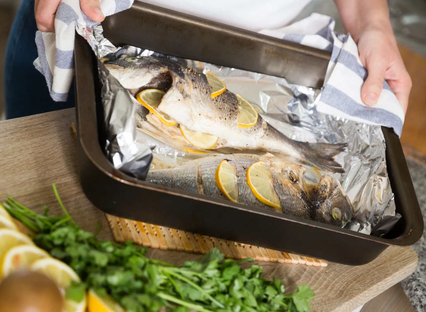 Dwie ryby ułożone w brytfannie wyłożonej folią aluminiową, przełożone plasterkami cytryny, trzymane przez osobę w białej koszulce. Na pierwszym planie świeża natka pietruszki oraz deska do krojenia.