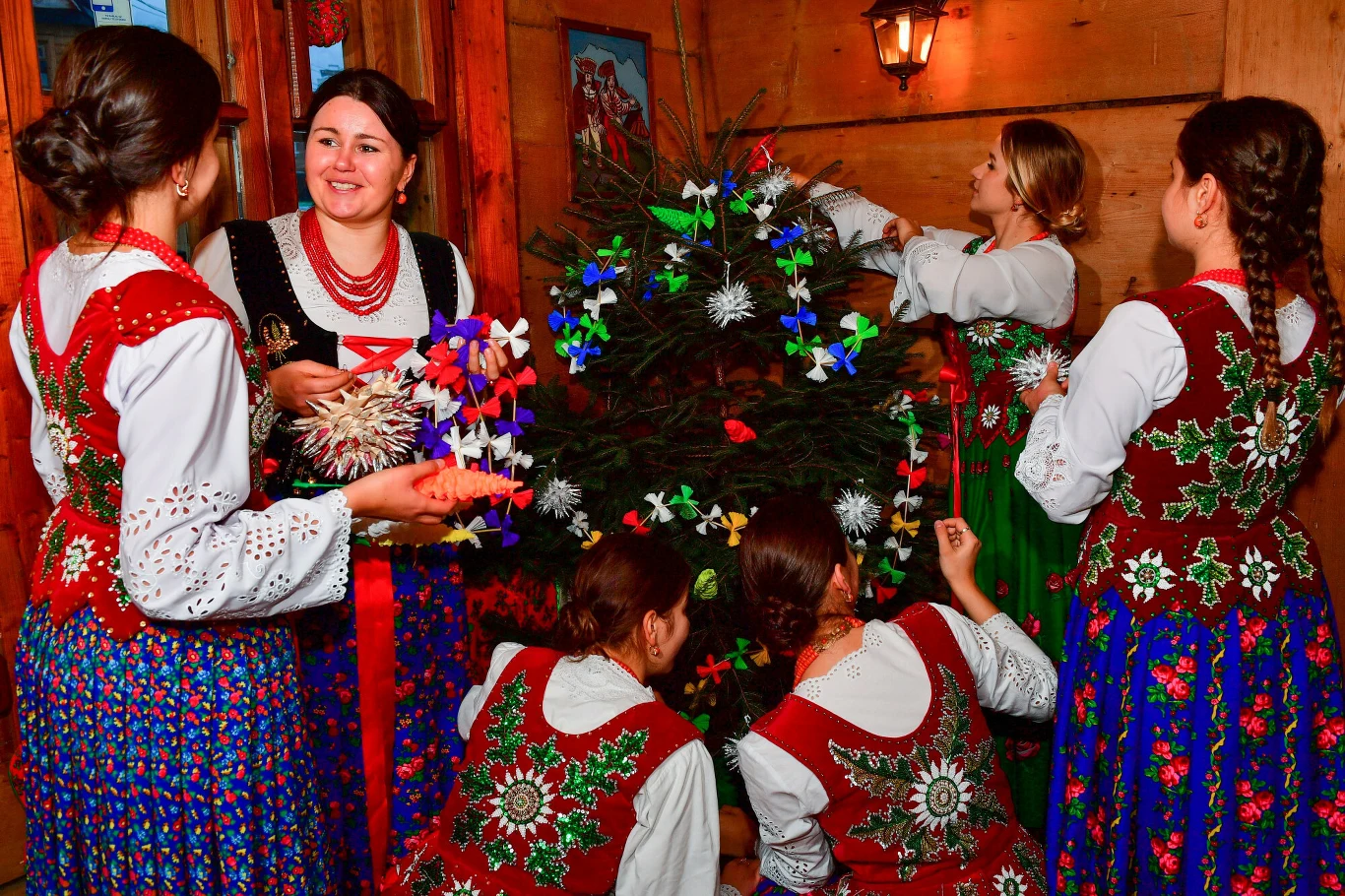 Grupa kobiet w tradycyjnych, kolorowych strojach ludowych dekoruje choinkę różnobarwnymi, ręcznie robionymi ozdobami w drewnianym wnętrzu, tworząc świąteczną, radosną atmosferę.