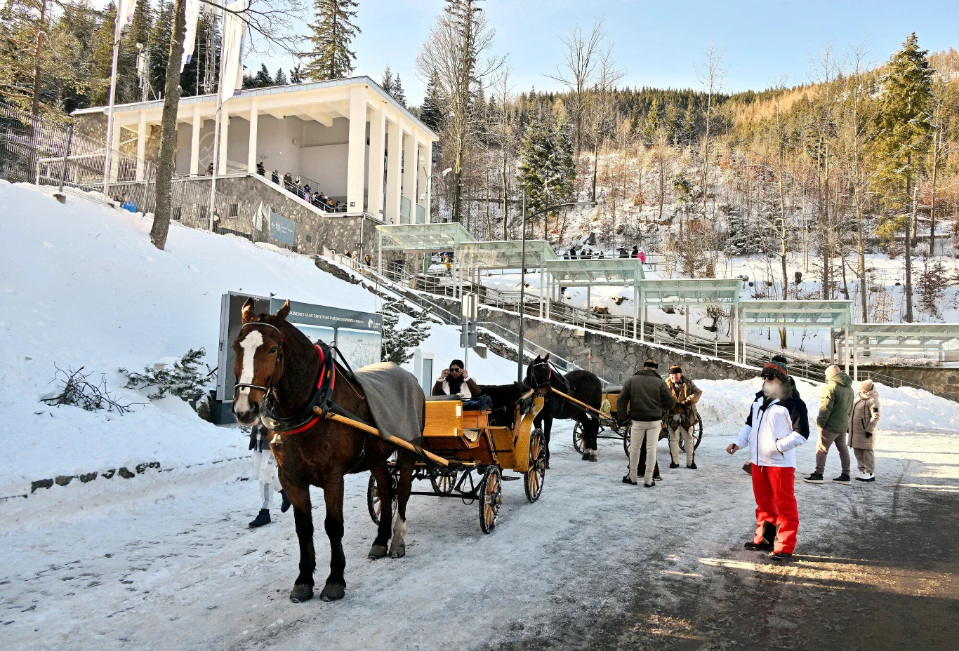Górski krajobraz zimowy z saniami zaprzężonymi w konie oraz ludźmi w ciepłych ubraniach. W tle biały budynek z kolumnadą, na zboczu lasu, widoczne alejki oraz grupy turystów. Dominują śnieg, drzewa iglaste i spokojna atmosfera wypoczynku.