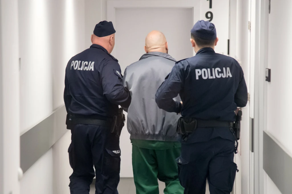 Sąd utrzymał wyrok ws. Krystiana W. ps. Krystek Dwóch funkcjonariuszy policji w mundurach prowadzi mężczyznę o łysiejących włosach w zielonych spodniach przez jasny korytarz, kierując się w stronę zamkniętych drzwi.