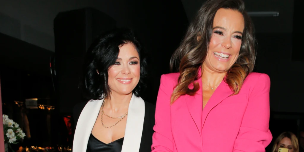 Katarzyna Cichopek i Anna Mucha Dwie kobiety ubrane elegancko, jedna w czarnym stroju z białą marynarką, druga w różowej marynarce, obie uśmiechnięte, stoją obok siebie na tle ciemnego wnętrza.