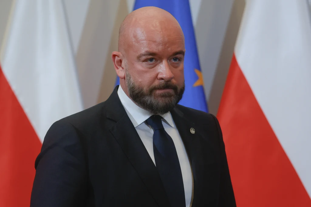 Prezydent Wrocławia Jacek Sutryk Mężczyzna w garniturze i krawacie stoi na tle flag Polski i Unii Europejskiej.