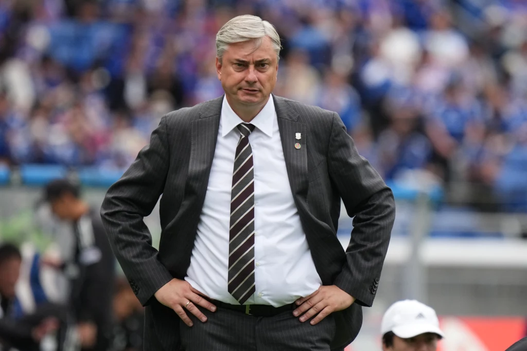 Maciej Skorża Mężczyzna w średnim wieku w eleganckim garniturze stoi na tle stadionu, z rękami opartymi o biodra, sprawia wrażenie skoncentrowanego i zamyślonego, w tle rozmyta publiczność i osoby zaangażowane w wydarzenie sportowe.