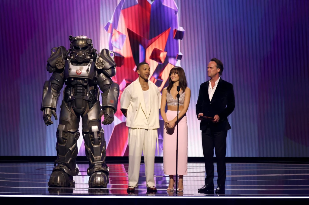 The Game Awards Cztery osoby stoją na scenie przed publicznością, z czego jedna występuje w dużym kostiumie robota, a pozostałe osoby ubrane są elegancko. Na tle sceny widać kolorową, abstrakcyjną grafikę.