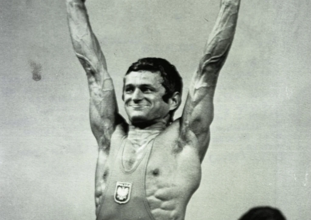 Zygmunt Smalcerz jest wielką legendą polskiej sztangi. To mistrz olimpijski i trzykrotny mistrz świata Mężczyzna w stroju sportowym z godłem Polski na piersi unosi ręce w geście zwycięstwa, uśmiechając się szeroko; wyraźnie widoczne umięśnienie i determinacja na twarzy.