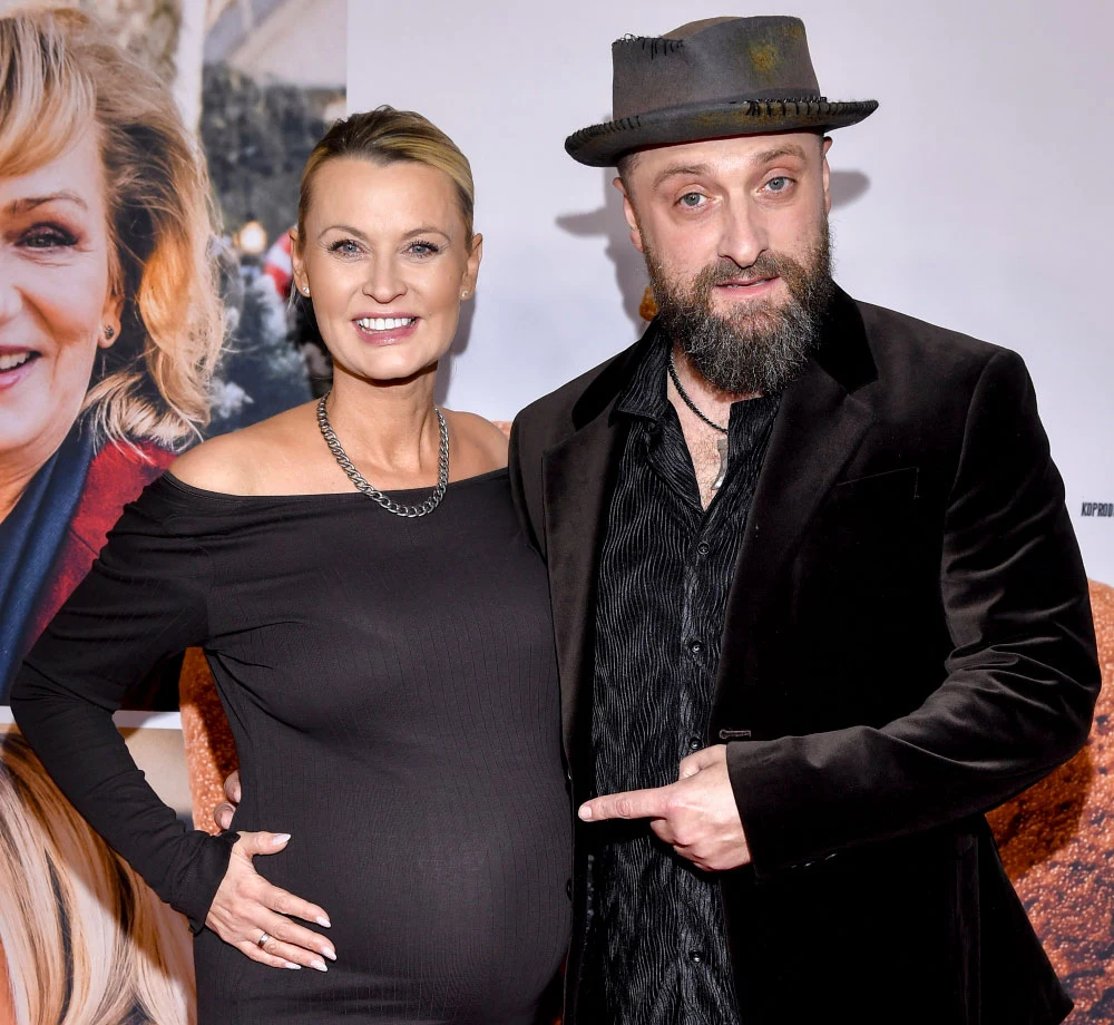 Monika Bradacz-Drapała i Łukasz Drapała ponownie zostali rodzicami Kobieta w eleganckiej czarnej sukni z widoczną ciążą stoi obok mężczyzny ubranego w czarny płaszcz i kapelusz; oboje uśmiechają się, a mężczyzna wskazuje dłonią na brzuch kobiety.