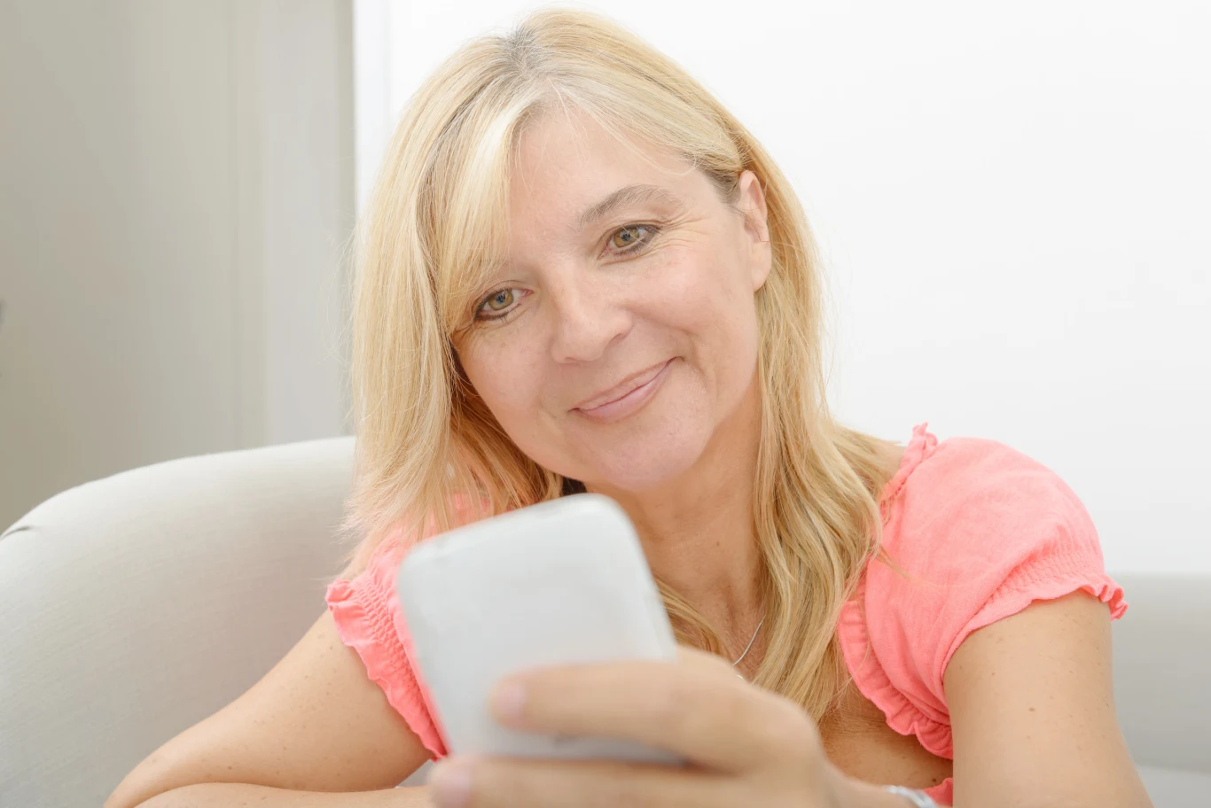 Naturalny peeling z kuchennych składników. Działa jak subtelny lifting Uśmiechnięta kobieta w średnim wieku siedzi na kanapie, trzymając w dłoni biały smartfon i patrząc na ekran z wyrazem zadowolenia.