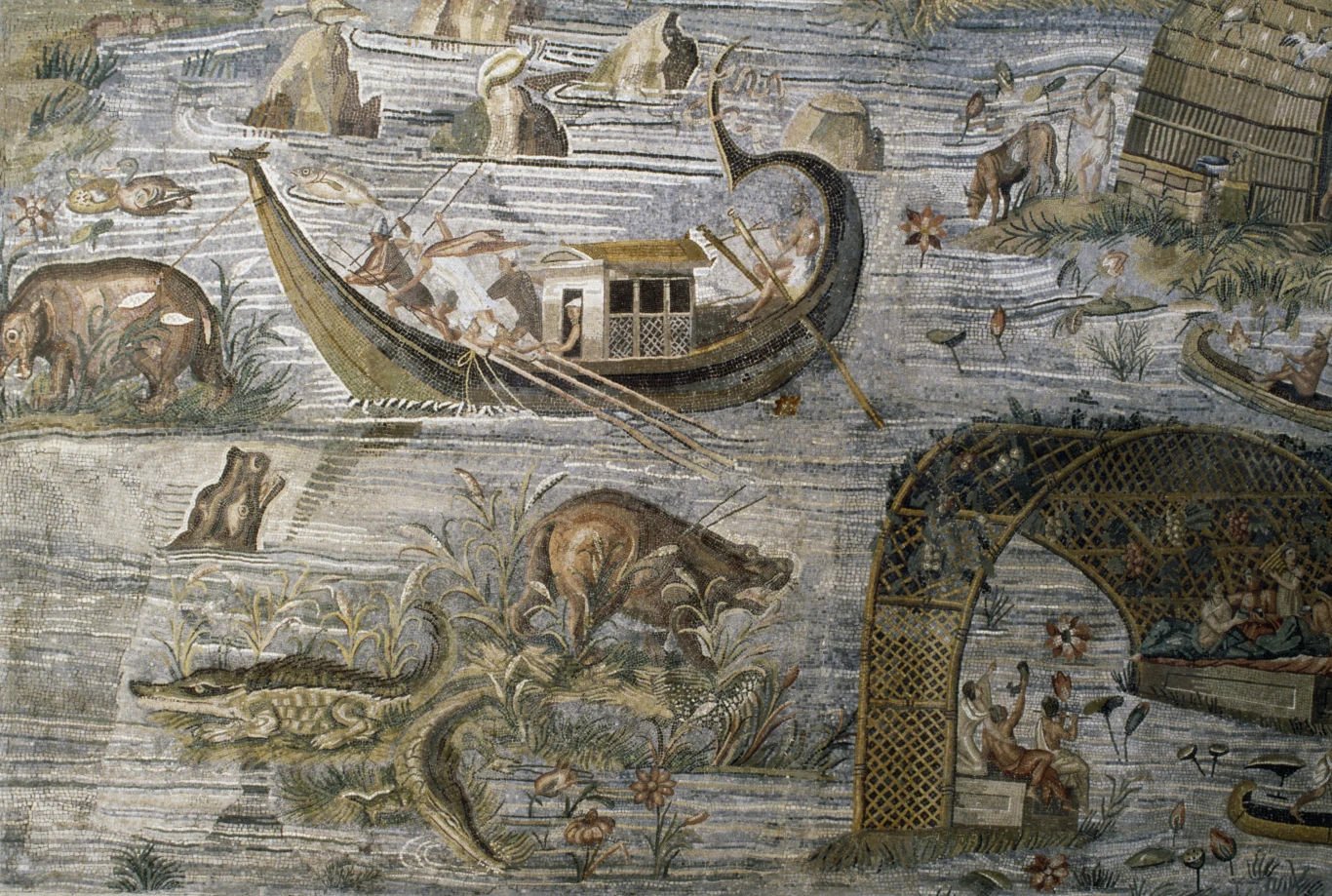 Łodzie i zwierzęta na Nilu. Detal z mozaiki nilotyckiej Kolorowa mozaika ukazująca scenę z życia nad rzeką. Na wodzie płyną łodzie z wioślarzami i pasażerami, na brzegu widoczne dzikie zwierzęta, w tym krokodyl i hipopotam. Po prawej stronie znajduje się zadaszona altana z odpoczywającymi ludźmi. W tle rzek...