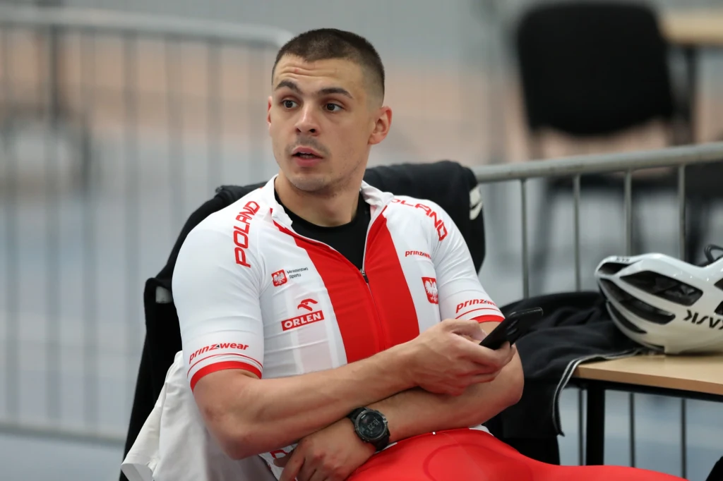 Patryk Rajkowski Młody mężczyzna w stroju kolarskim reprezentacji Polski siedzi przy stole, trzyma telefon w ręce i patrzy w bok. Na stole leży kask rowerowy, w tle widoczne są krzesła i barierki.
