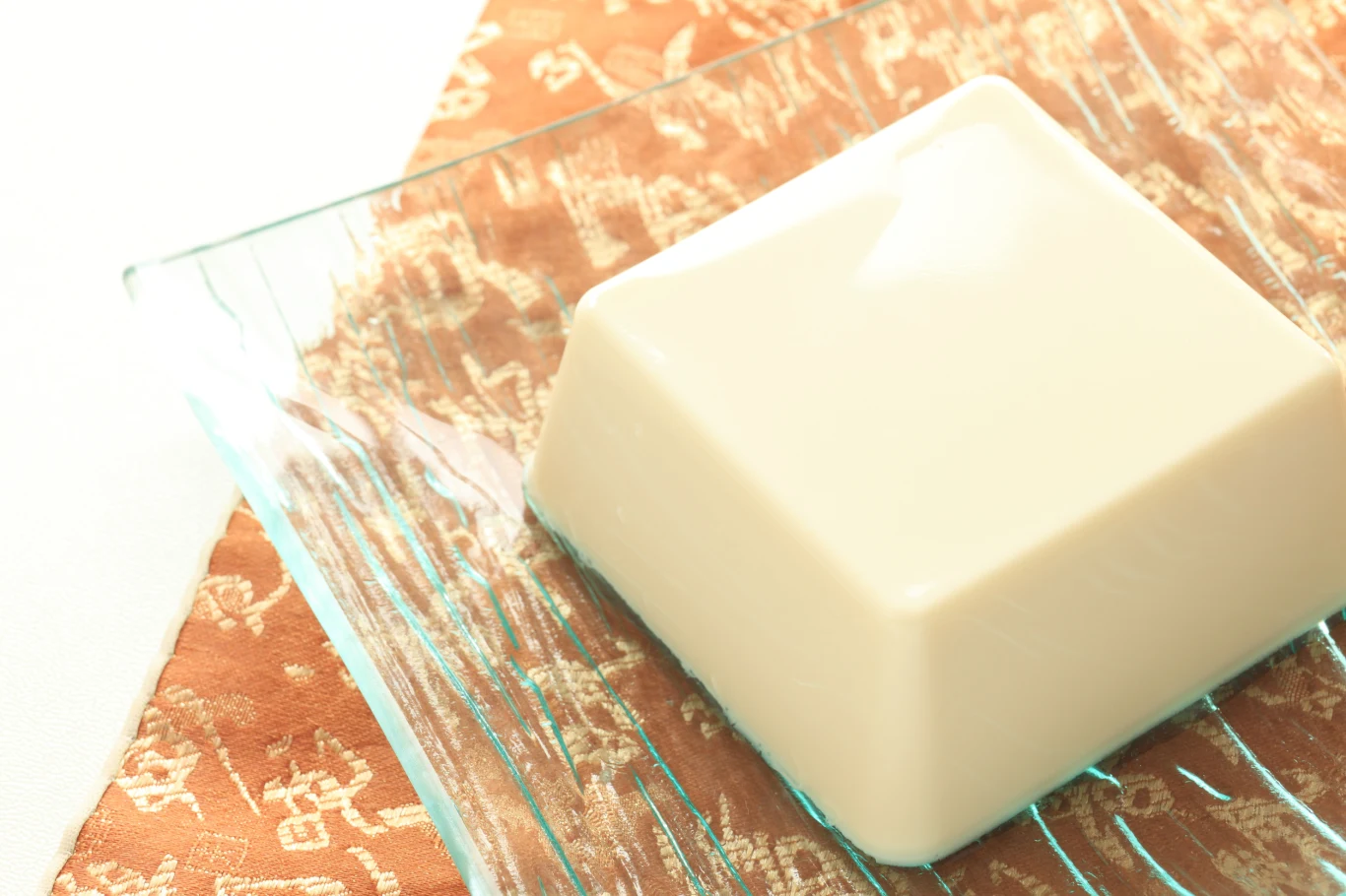 Domowe mydło, które poprawia komfort skóry już po tygodniu Kostka jasnego tofu umieszczona na przezroczystym, szklanym talerzu z fantazyjnymi liniami, leżącym na ozdobnej tkaninie w kolorze pomarańczowym z delikatnym wzorem.