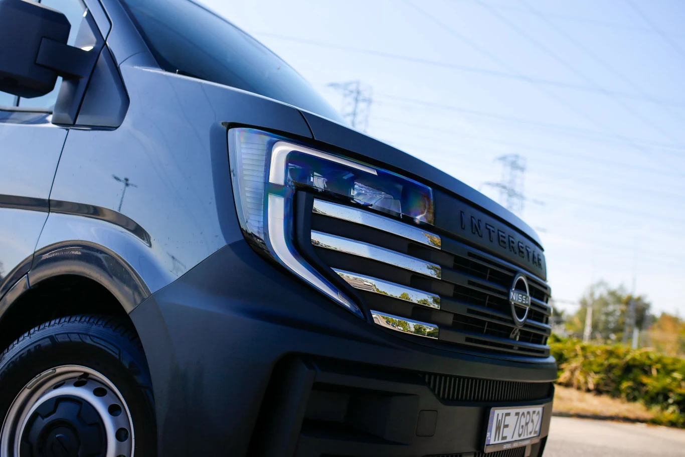 Nowoczesny przód szarego vana marki Nissan z wyraźnym grillem i dużym reflektorem, srebrne elementy podkreślające design pojazdu. W tle asfalt, zieleń i słupy wysokiego napięcia.