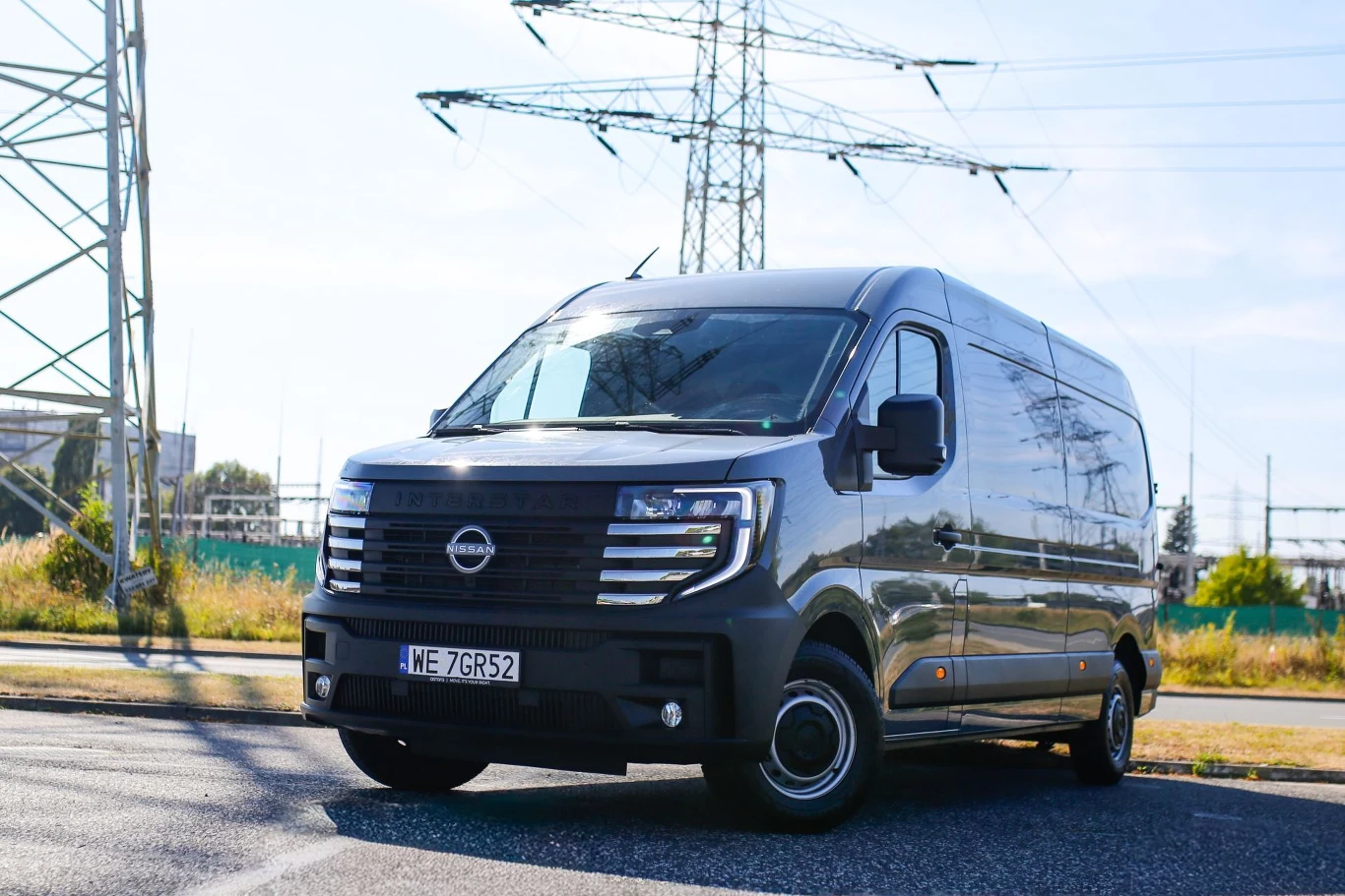 Nowoczesny czarny van marki Nissan ustawiony na ulicy w słoneczny dzień, w tle widoczne słupy energetyczne i zielone elementy krajobrazu miejskiego.