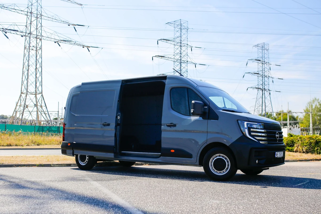 Duży ciemnoszary samochód dostawczy z otwartymi drzwiami bocznymi stoi na asfalcie, w tle widoczne są maszty wysokiego napięcia oraz zieleń i błękitne niebo.