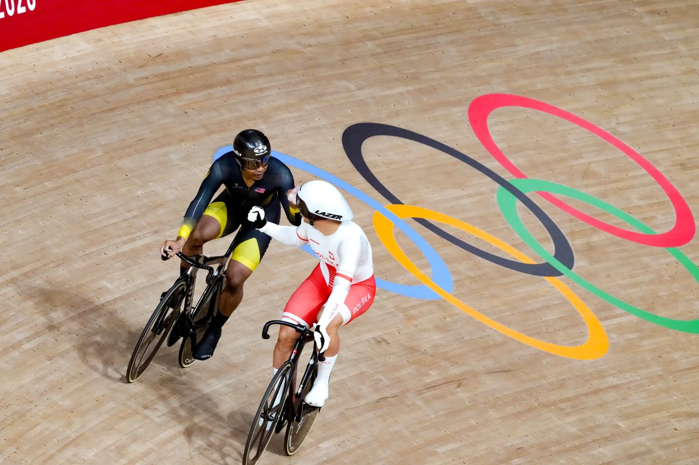 Dwóch kolarzy torowych rywalizujących na welodromie, jeden ubrany w biało-czerwony strój, drugi w czarno-żółto-niebieski. Jadą obok siebie na tle wyraźnych olimpijskich kół na drewnianym torze.