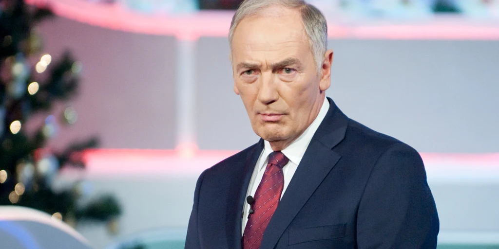 Karol Strasburger Elegancko ubrany mężczyzna w garniturze i czerwonym krawacie, stojący w studiu telewizyjnym, w tle świąteczna dekoracja z choinką i różowym oświetleniem.