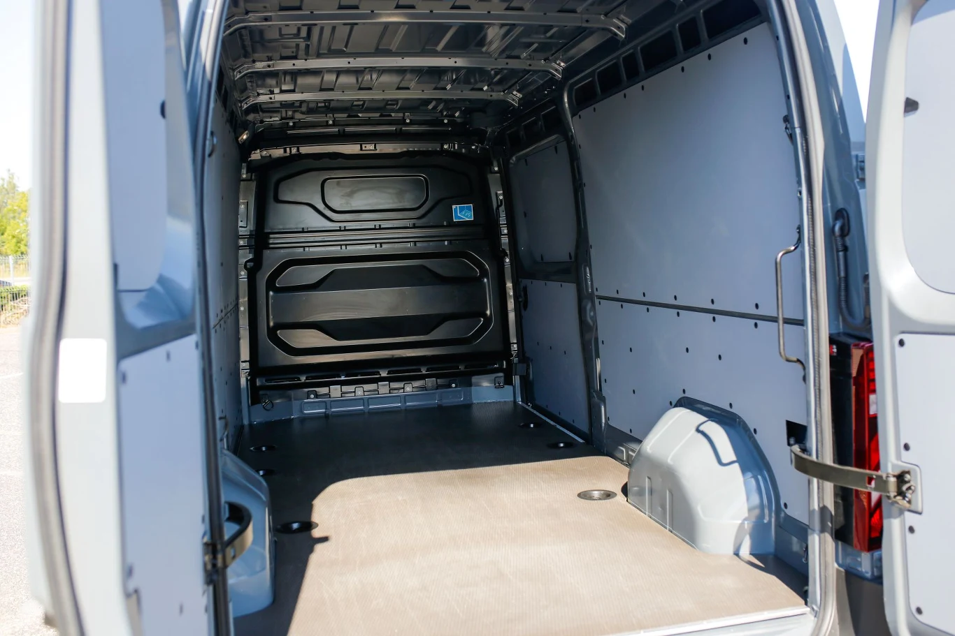 Puste wnętrze samochodu dostawczego z widoczną metalową podłogą oraz ścianami bocznymi, bez obecności towaru lub osób. W drzwiach bocznych widoczna klamka, wnętrze oświetlone światłem dziennym.