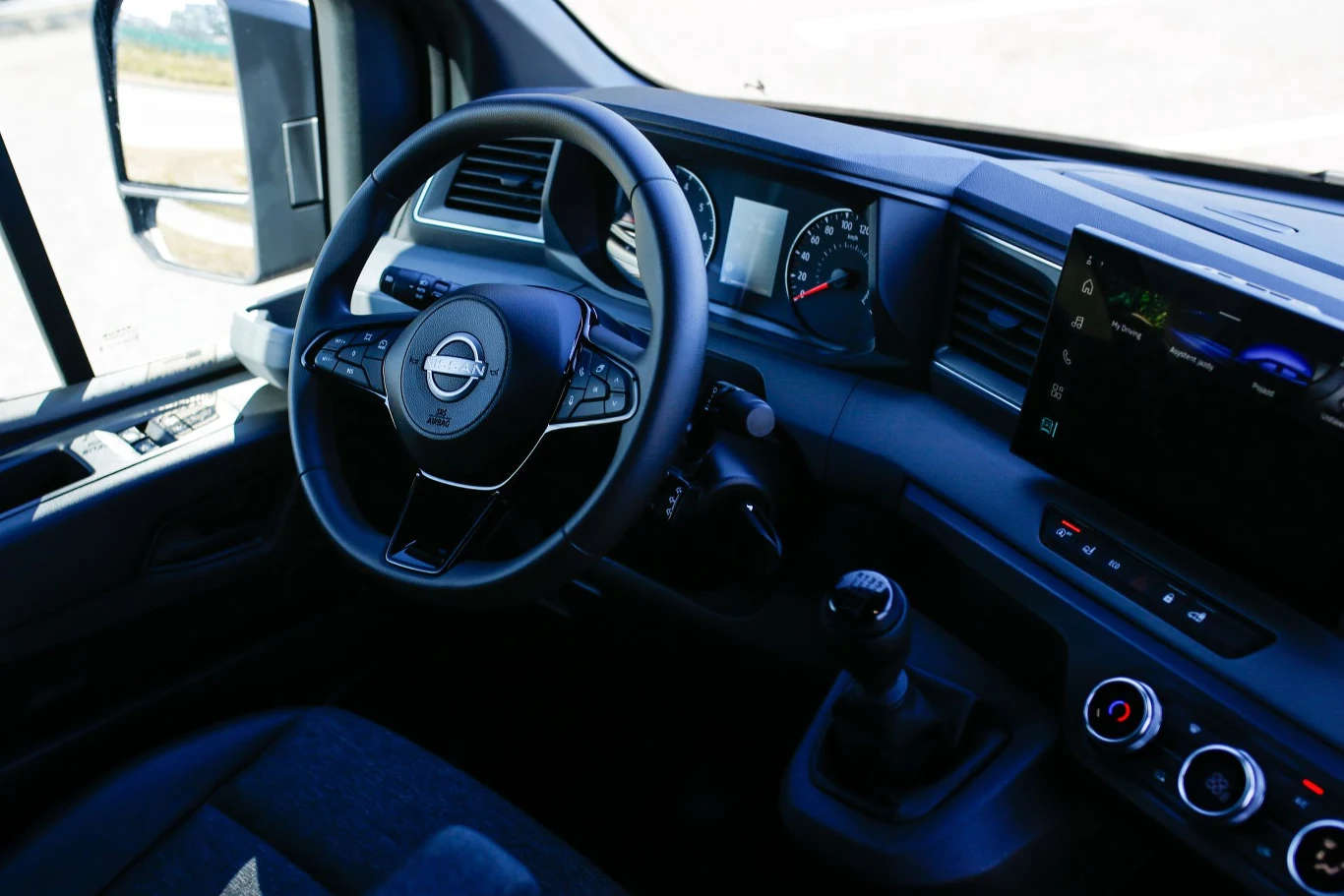 Nowoczesny kokpit samochodu dostawczego z widoczną kierownicą, zegarami na desce rozdzielczej, manualną skrzynią biegów, wyświetlaczem multimedialnym i panelami sterowania klimatyzacją.