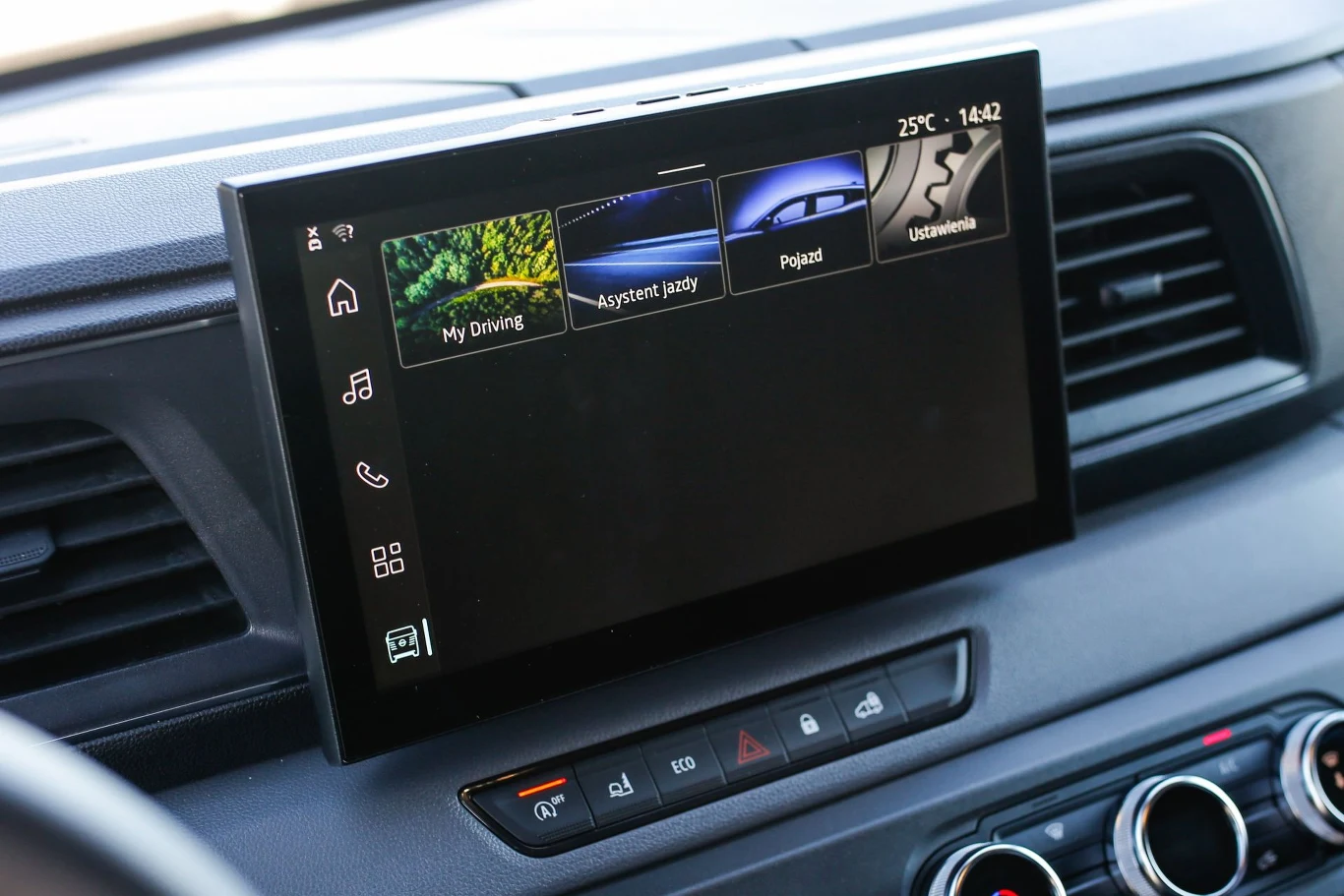 Dotykowy wyświetlacz systemu multimedialnego zamontowany na desce rozdzielczej samochodu, pokazujący różne aplikacje i opcje sterowania, u dołu widoczny panel z przyciskami i pokrętłami klimatyzacji.