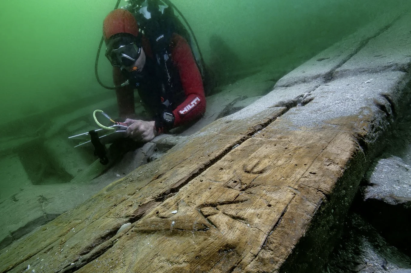 Greckie "graffiti" znalezione na jednym z elementów statku, datowane na pierwszą połowę I wieku n.e. Nurek w specjalistycznym stroju bada zatopiony drewniany wrak statku znajdujący się na dnie zbiornika wodnego, wykonując pomiary narzędziami naukowymi.