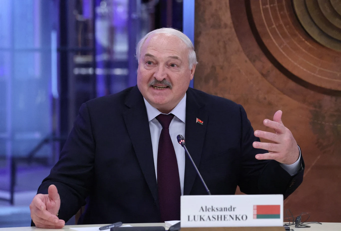 Łukaszenko chce przygotować Białoruś do potencjalnej wojny Mężczyzna w garniturze siedzący przy stole z mikrofonem podczas oficjalnego spotkania, przed nim znajduje się tabliczka z imieniem i nazwiskiem oraz flagą państwa.
