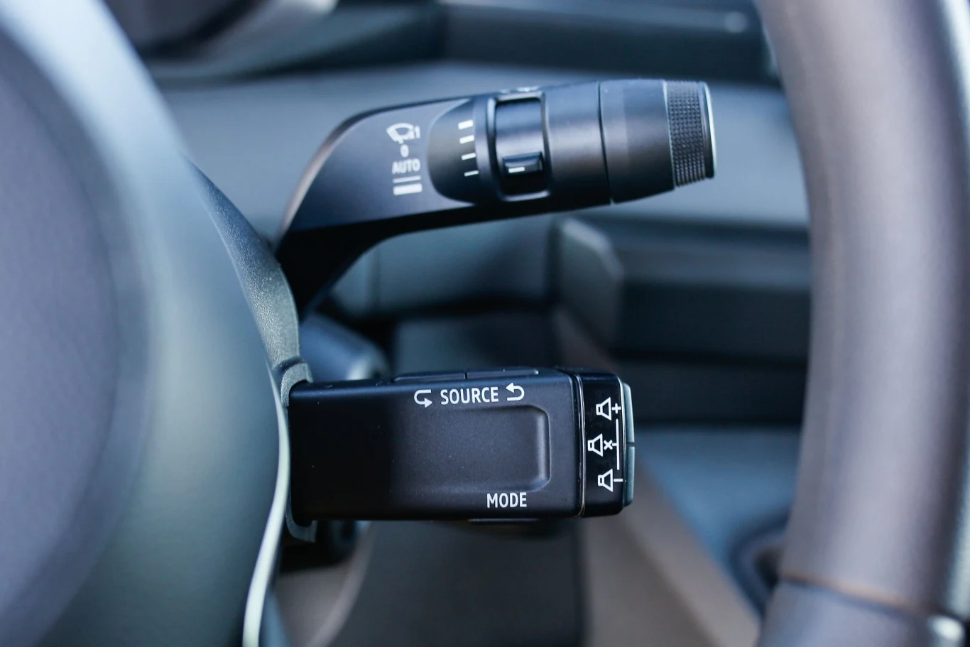 Dźwignia zmiany trybów oraz sterowania funkcjami na kierownicy samochodu, widoczne są symbole wskazujące na konkretne funkcje, a także fragment kolumny kierowniczej i części wnętrza auta.