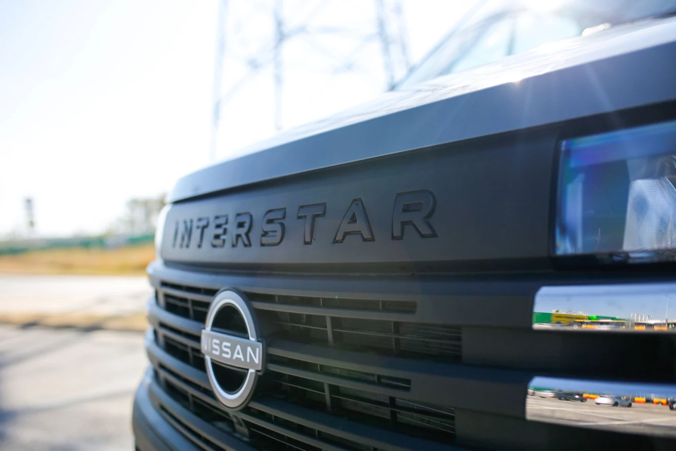 Zbliżenie na przód samochodu dostawczego marki Nissan Interstar z widocznym logotypem i maską pojazdu, w tle rozmyte elementy infrastruktury.