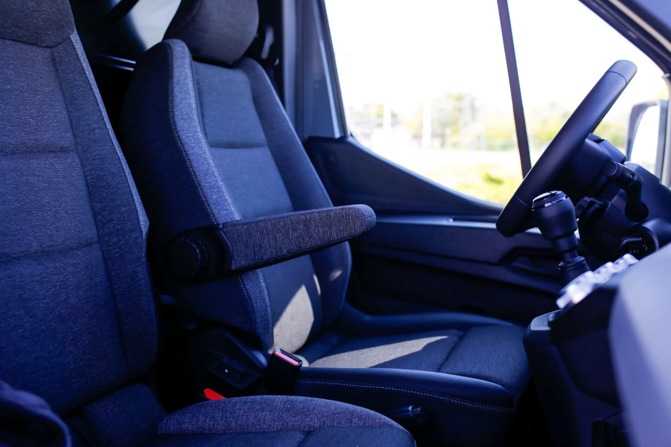 Przednie fotele samochodu wykonane z ciemnego materiału, między siedzeniami znajduje się podłokietnik, po prawej widoczna jest kierownica i fragment deski rozdzielczej, tło rozmyte pokazuje światło słoneczne wpadające przez boczne szyby.