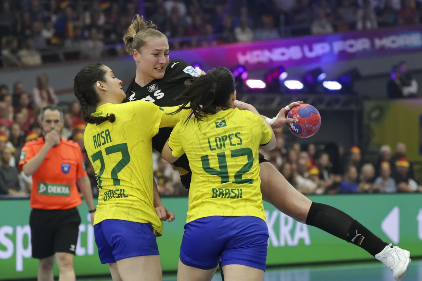 Intensywna akcja podczas meczu piłki ręcznej, zawodniczka w czarnym stroju próbuje rzucić piłkę, będąc blokowaną przez dwie rywalki z brazylijskiej drużyny w żółto-niebieskich strojach. W tle publiczność oraz sędzia obserwujący grę.