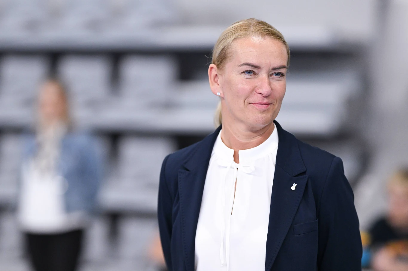 Kobieta w eleganckim granatowym żakiecie i białej bluzce stoi na pierwszym planie w rozmytym pomieszczeniu z szarym tłem, delikatnie uśmiechnięta i patrzy w bok.