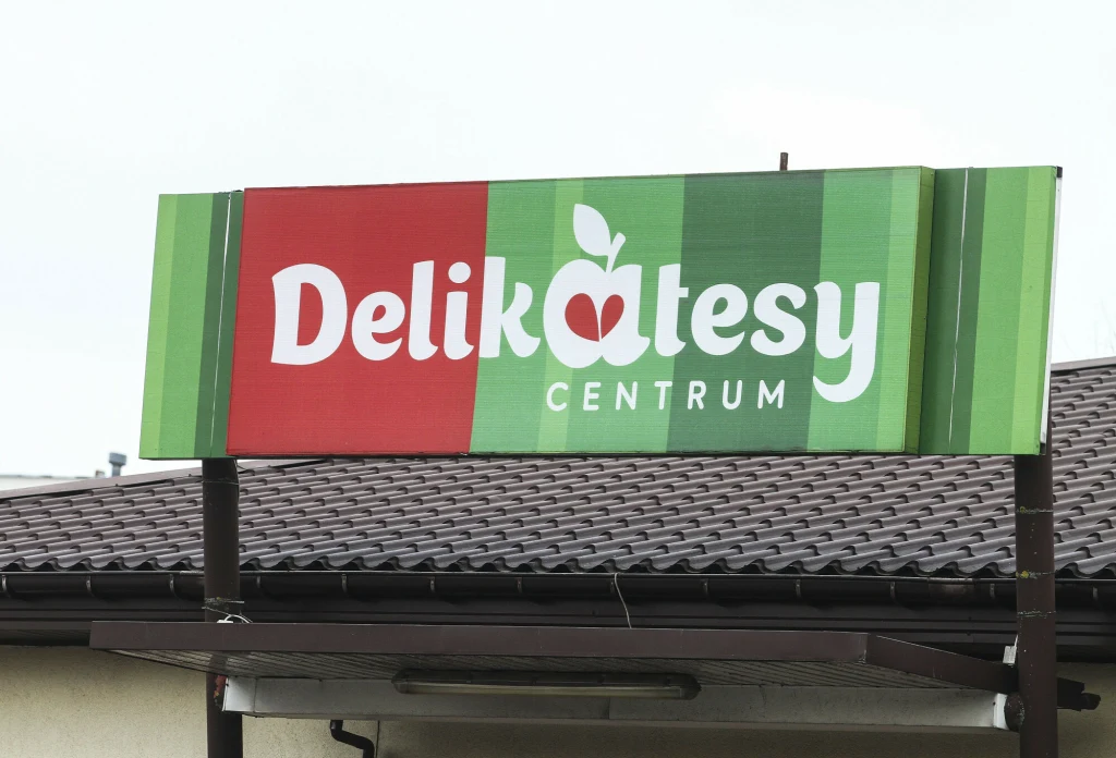 Szyld sklepu Delikatesy Centrum z kolorowym napisem i motywem jabłka na tle dachówki budynku.