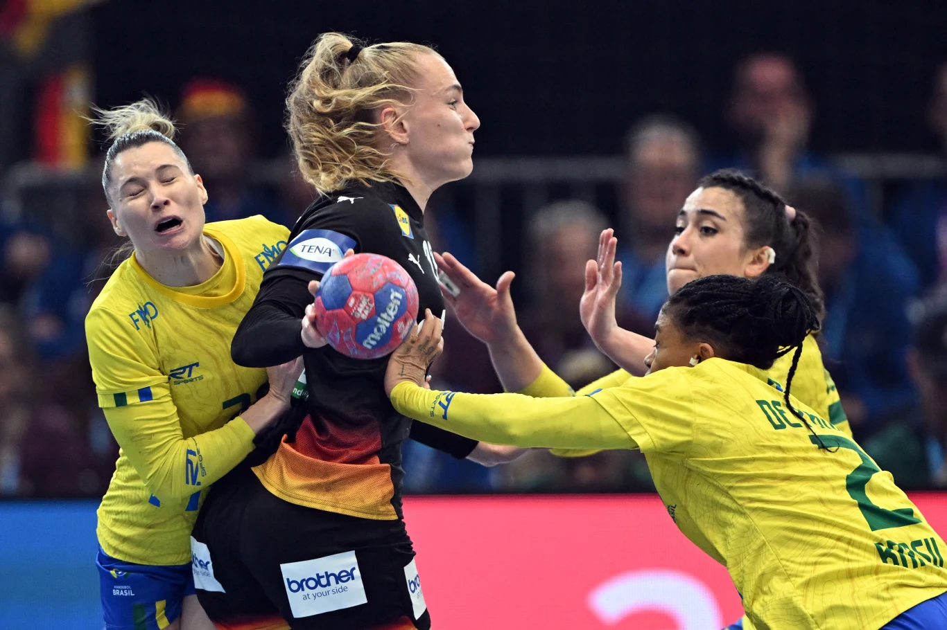 Intensywna walka o piłkę podczas meczu piłki ręcznej, cztery zawodniczki z dwóch drużyn są mocno zaangażowane w akcję, jedna zawodniczka w czarnym stroju ściskając piłkę próbuje przedrzeć się przez rywalki w żółtych strojach, w tle widoczni są kibice.