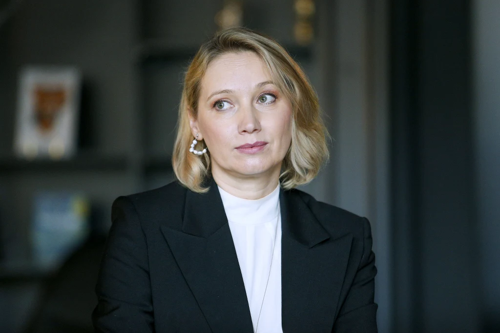 Joanna Orleańska Kobieta o blond włosach w eleganckiej czarnej marynarce i białej bluzce, siedząca w pomieszczeniu z rozmytym, stonowanym tłem. Spogląda w bok, wyrażając zamyślenie lub refleksję.