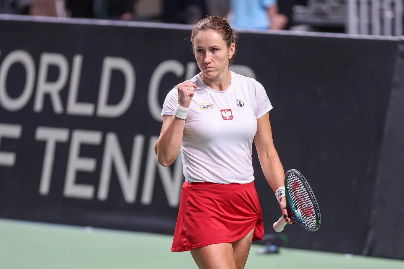 Kobieta ubrana w biały podkoszulek i czerwoną spódniczkę w trakcie meczu tenisowego, trzyma rakietę tenisową w prawej ręce i wykonuje gest triumfu zaciśniętą pięścią, w tle napis na ścianie kortu.