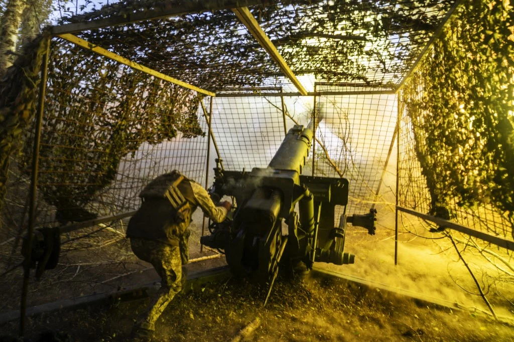 Żołnierz w mundurze obsługujący ciężką armatę polową ukrytą pod siatką maskującą, wśród wzbijającego się dymu i rozbłysku ognia podczas wystrzału.