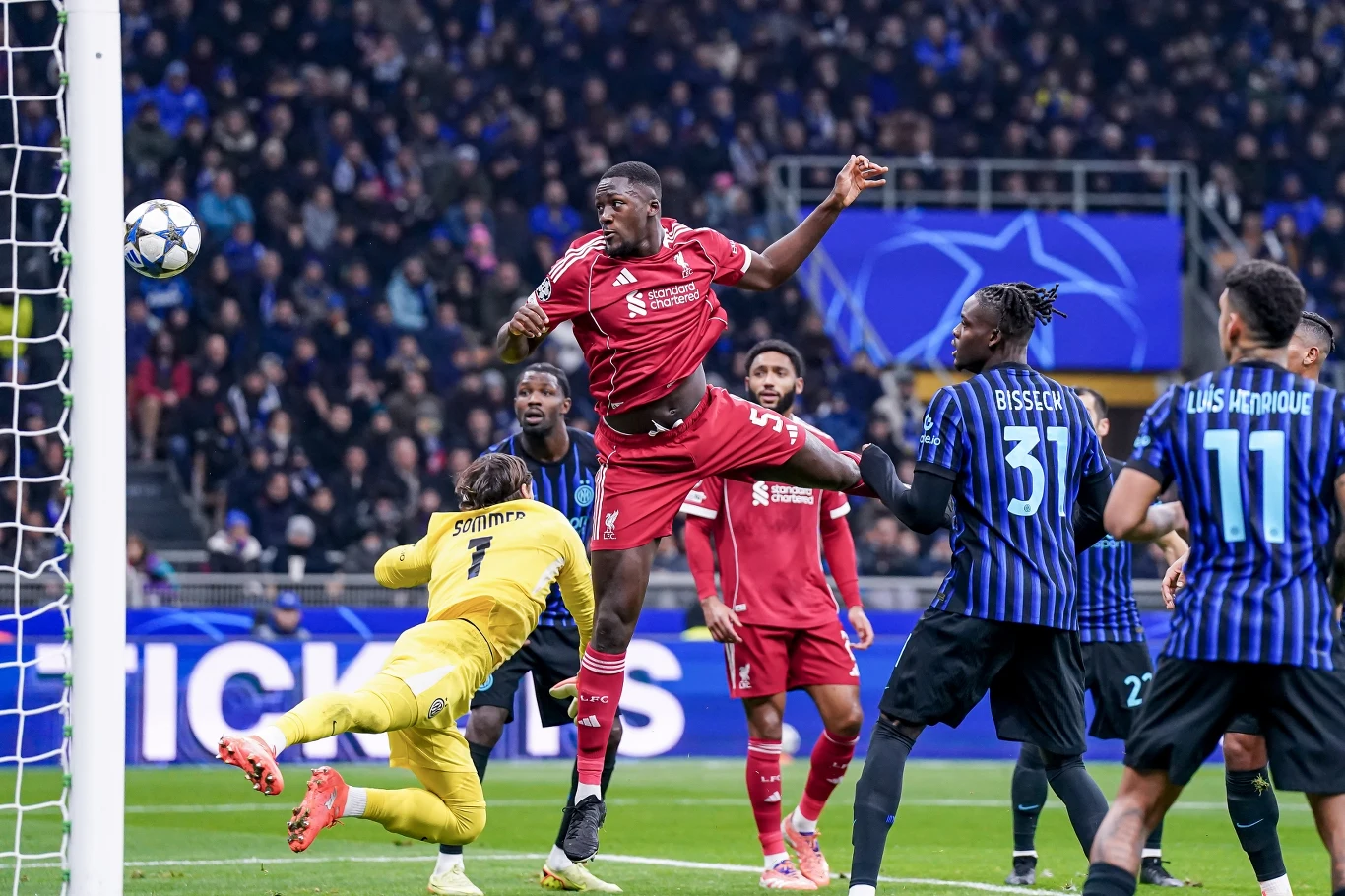 Dynamiczna akcja podczas meczu piłki nożnej, zawodnik w czerwonym stroju wyskakuje do piłki w polu karnym, bramkarz w żółtym stroju próbuje interweniować, otoczony piłkarzami w niebiesko-czarnych koszulkach, trybuny pełne kibiców w tle.