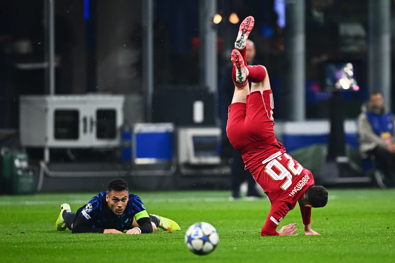 Dwóch piłkarzy w trakcie dynamicznej akcji meczowej, jeden z nich upada na murawę wykonując przewrót, drugi leży na ziemi patrząc w jego stronę, wokół widać zieloną nawierzchnię boiska oraz rozmazane sylwetki i sprzęt przy linii bocznej.