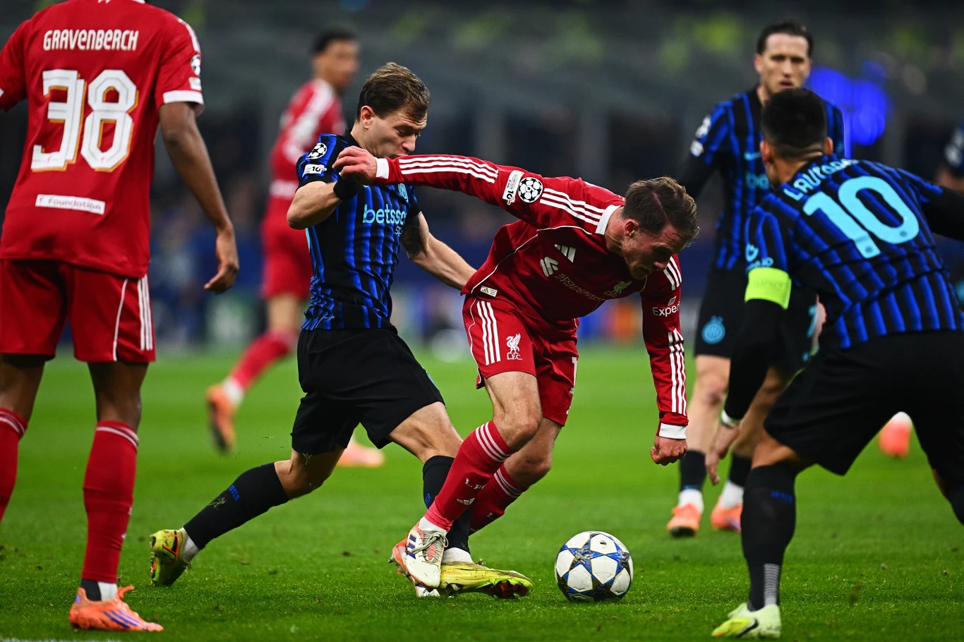 Piłkarz w czerwonym stroju próbuje opanować piłkę, otoczony przez zawodników drużyny przeciwnej w niebiesko-czarnych koszulkach. Jeden z rywali próbuje odebrać mu piłkę, pozostali zawodnicy obserwują sytuację na boisku.