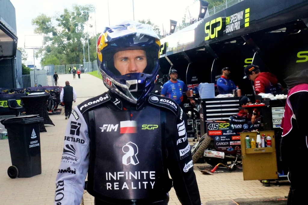 Zawodnik żużlowy w czarno-białym kombinezonie z kaskiem na głowie stoi na torze żużlowym w otoczeniu mechaników i sprzętu przygotowanego do wyścigu. W tle znajdują się stanowiska serwisowe oraz pracownicy zespołów.