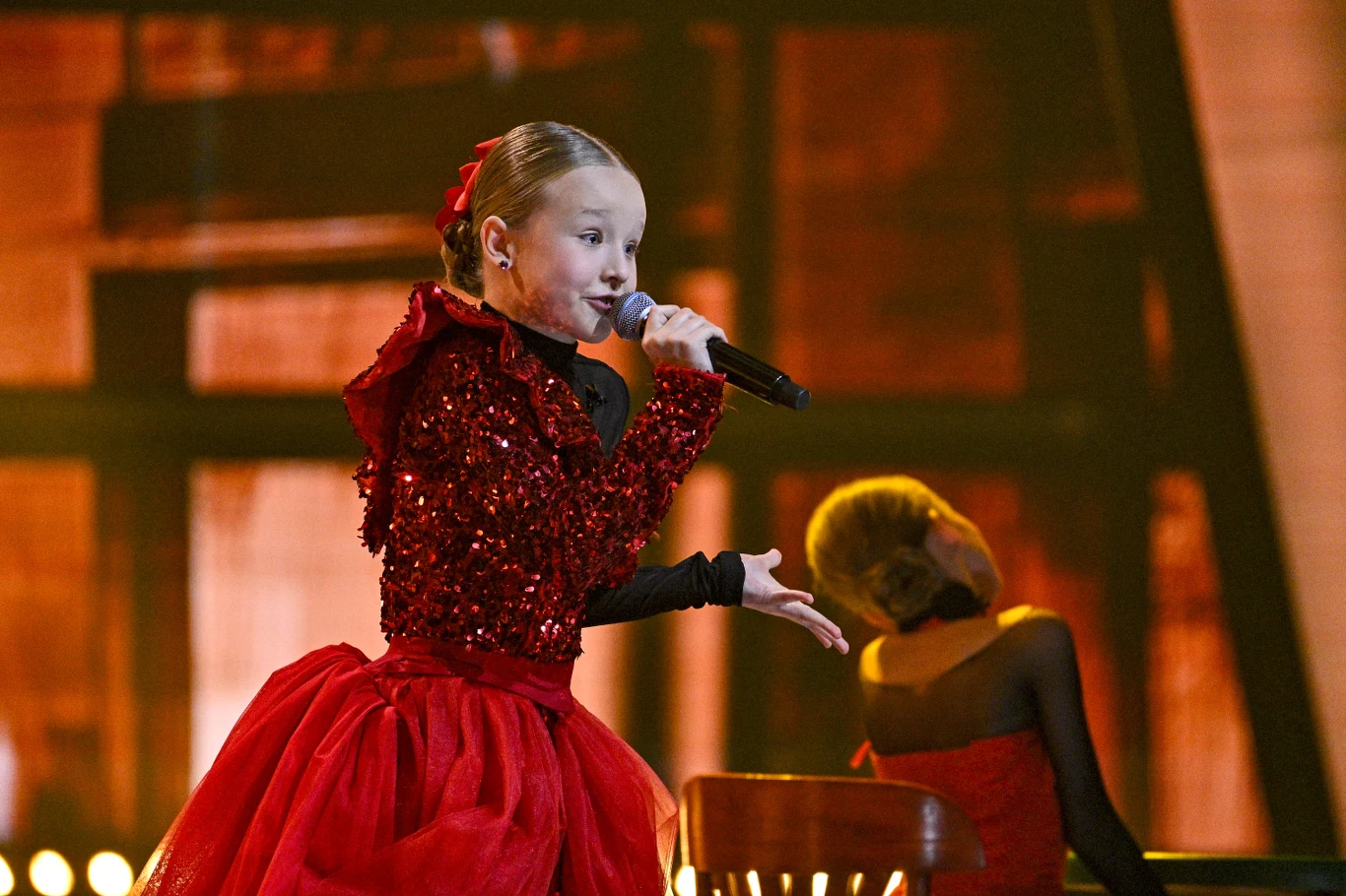 Dziewczynka w czerwonej, połyskującej sukni śpiewająca do mikrofonu na scenie, w tle siedzi inna dziewczynka ubrana w podobną stylizację.