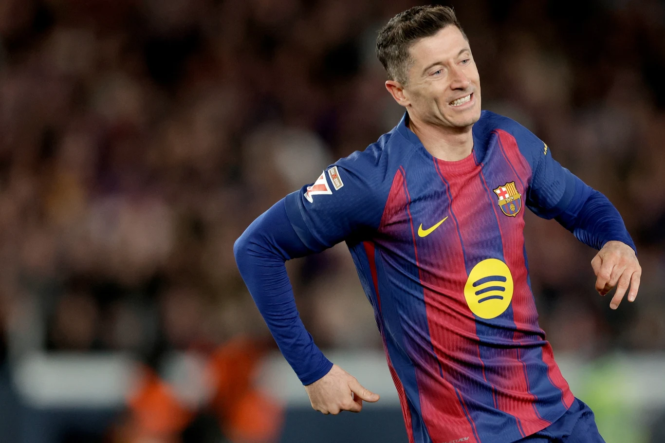 Robert Lewandowski Piłkarz drużyny FC Barcelona w klubowym stroju sportowym biegnie po boisku podczas meczu, w tle rozmazana publiczność stadionu.