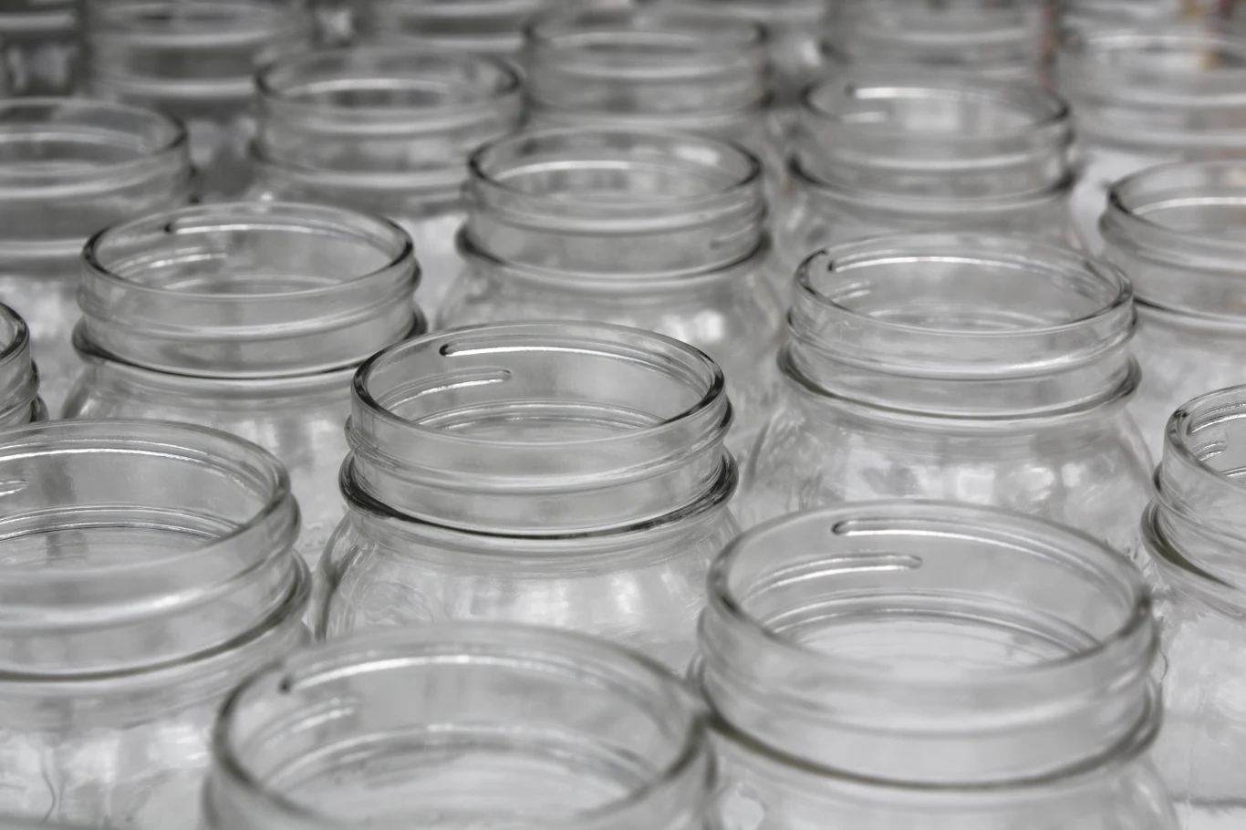 Rzędy pustych, przezroczystych szklanych słoików ustawionych blisko siebie tworzą regularny wzór. Brak zawartości i etykiet, szkło jest czyste i nieuszkodzone.