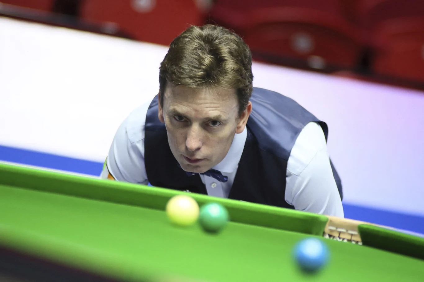 Snookerzysta w kamizelce i muchą koncentruje się nad stołem bilardowym, na którym widoczne są kolorowe bile.