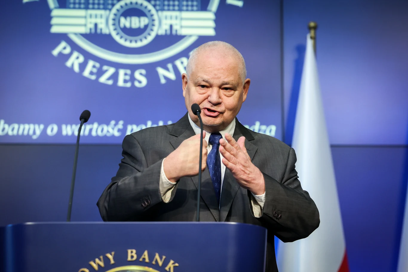 W kolejnym roku kończą się kadencje trzech członków zarządu NBP. Dla prezesa Adama Glapińskiego może to oznaczać kłopoty - pisze money.pl Mężczyzna w garniturze stoi za mównicą z logo Narodowego Banku Polskiego, gestykuluje podczas przemówienia, w tle widoczna flaga Polski oraz emblemat NBP.