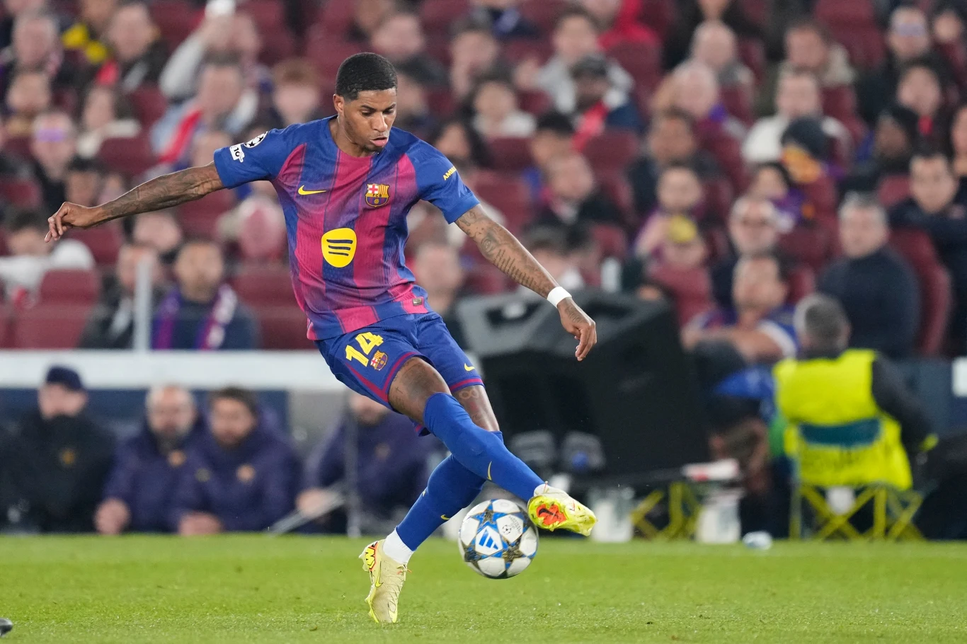 Piłkarz w stroju FC Barcelony kopie piłkę na stadionie podczas meczu, wokół widoczna publiczność na trybunach oraz kilku członków obsługi technicznej przy murawie.