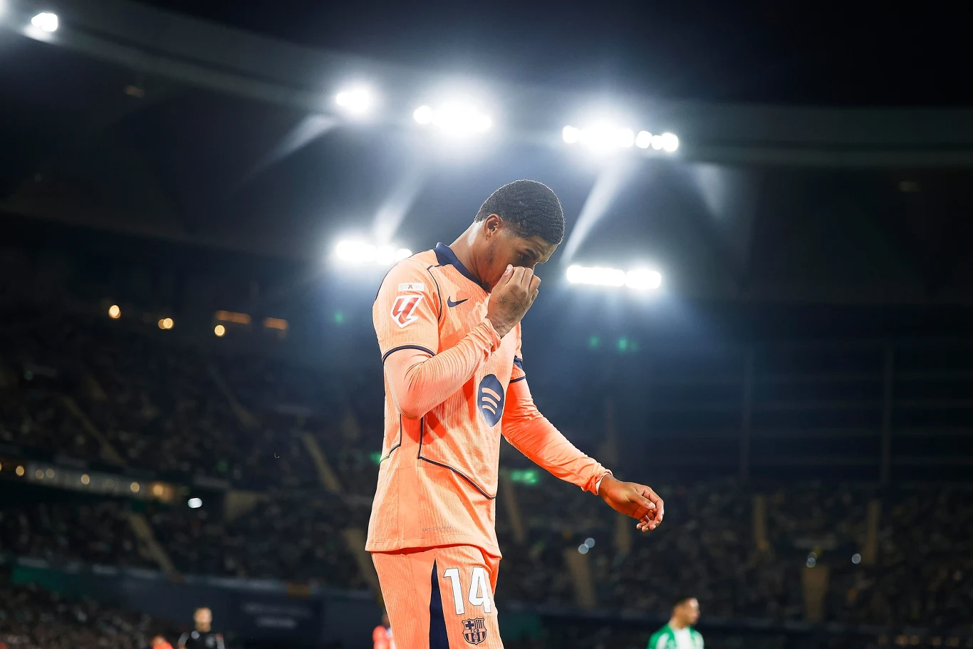 Piłkarz w pomarańczowym stroju drużynowym stoi na boisku pod mocnymi reflektorami, trzymając dłoń przy twarzy w geście smutku lub rozczarowania, w tle widać rozmyte sylwetki innych zawodników i trybuny stadionu.