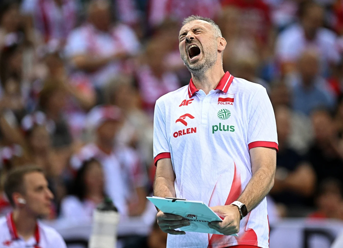 Trener siatkarski w białej koszulce z logotypami sponsorów energicznie reaguje na wydarzenia podczas meczu, trzymając notatki w dłoni, w tle widoczni kibice ubrane w biało-czerwone barwy.