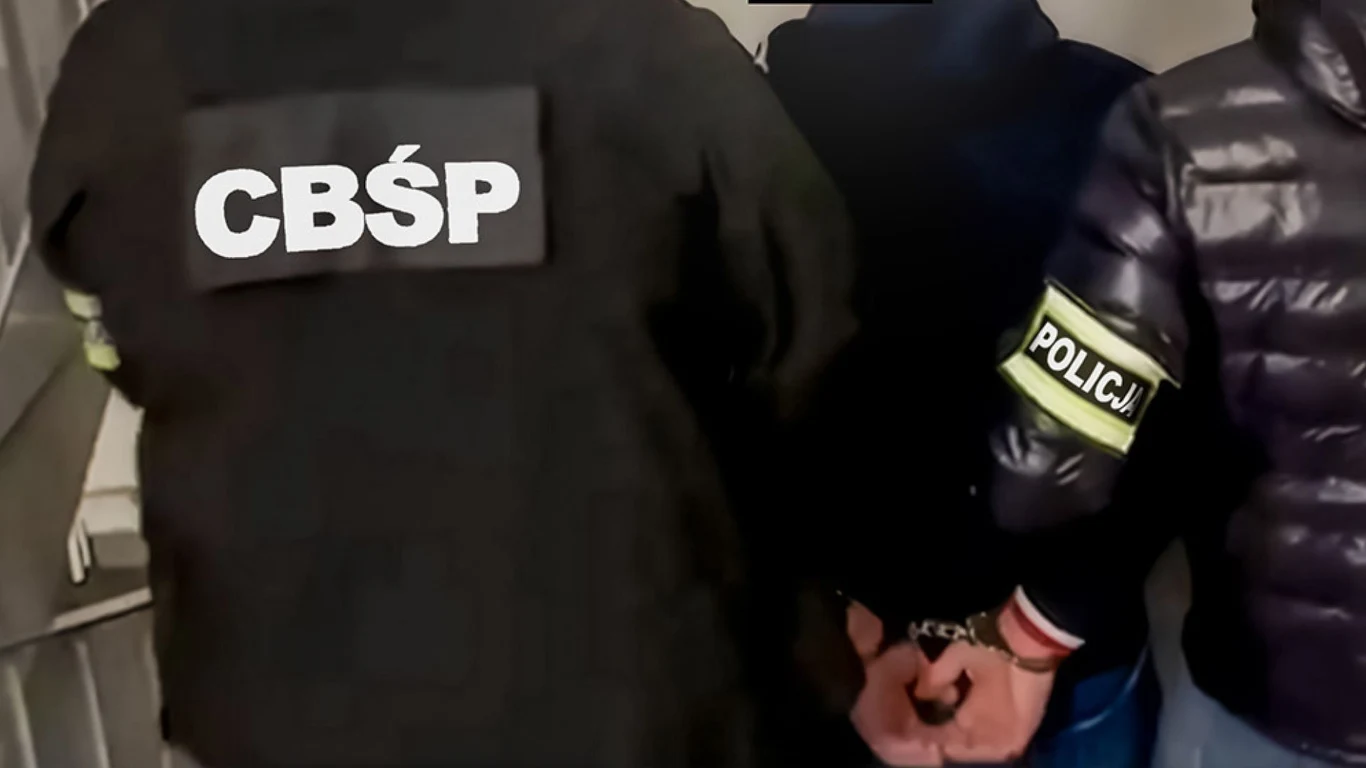 Rozbito zorganizowana grupę przestępczą. Legalizowali pobyt cudzoziemców Funkcjonariusze CBŚP i Policji prowadzą osobę w kajdankach, ubraną w ciemną kurtkę, przez korytarz lub klatkę schodową. Na ramionach funkcjonariuszy widoczne są oznaczenia służb.