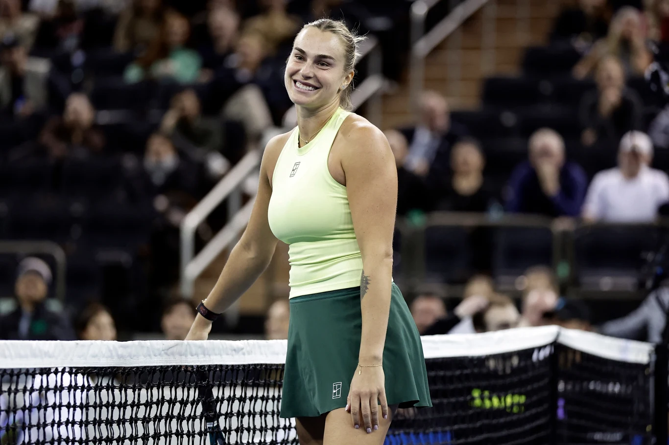 Aryna Sabalenka Uśmiechnięta zawodniczka tenisa na korcie, ubrana w jasnożółty top i zieloną spódniczkę, opierająca się o siatkę. W tle rozmazana publiczność na trybunach.