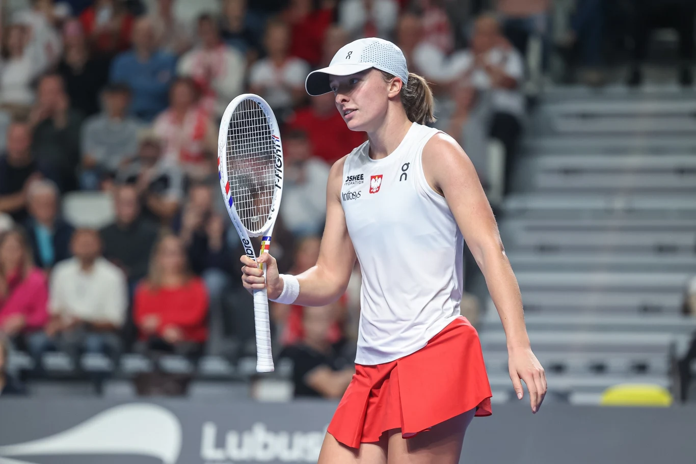 Iga Świątek Tenisistka w białej czapce i stroju sportowym z rakietą do tenisa w dłoni znajduje się na korcie podczas meczu, w tle widownia.